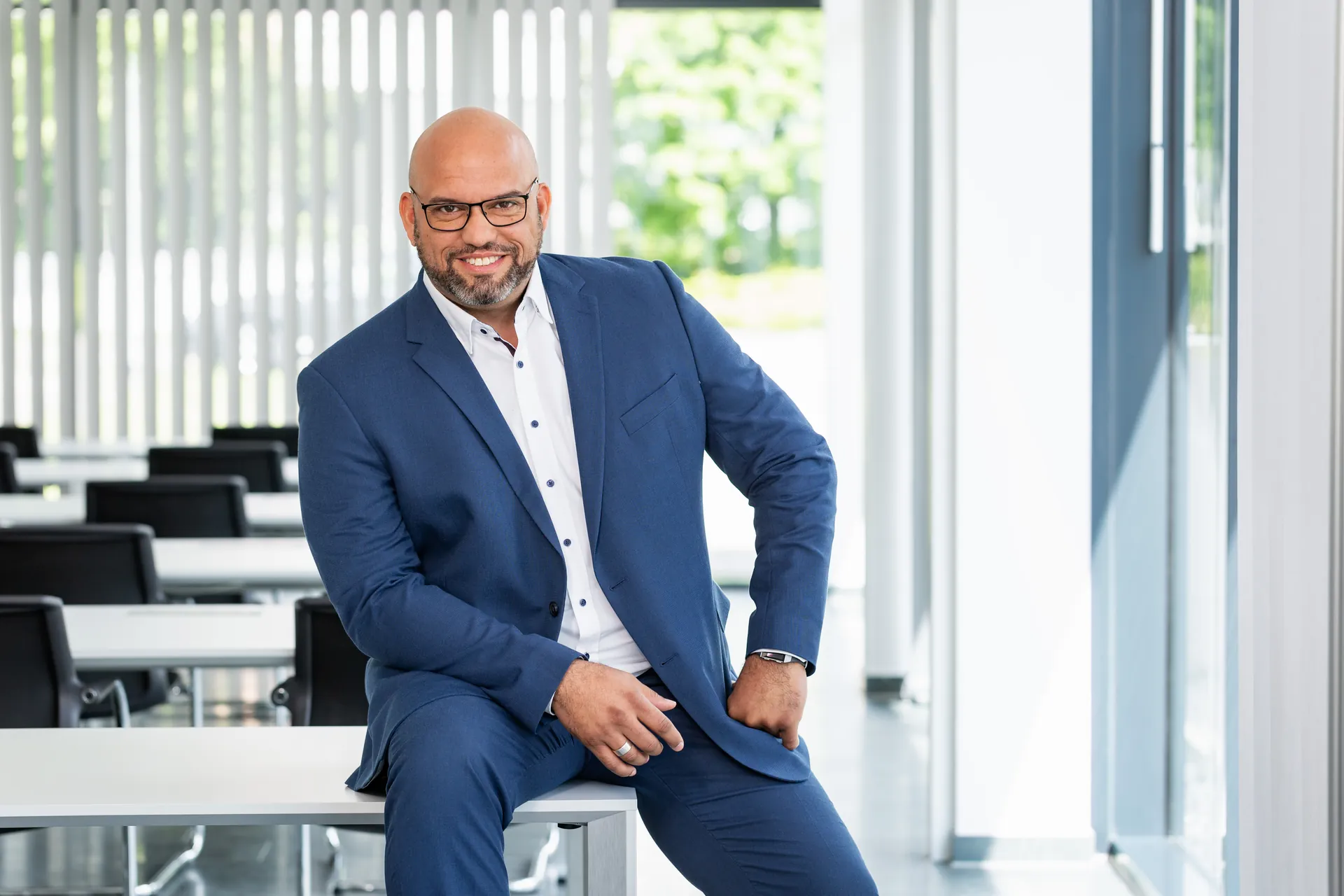 Pierre Mc Conner im blauen Anzug, auf einem Schreibtisch in einem hellen Büro sitzend